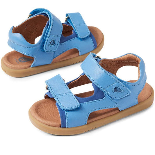 The Bobux IW Iwalk Strand Sandal in Azure Blue features toddler-friendly adjustable Velcro straps, tan soles, open toes and heels, and washable material, all displayed on a white background.