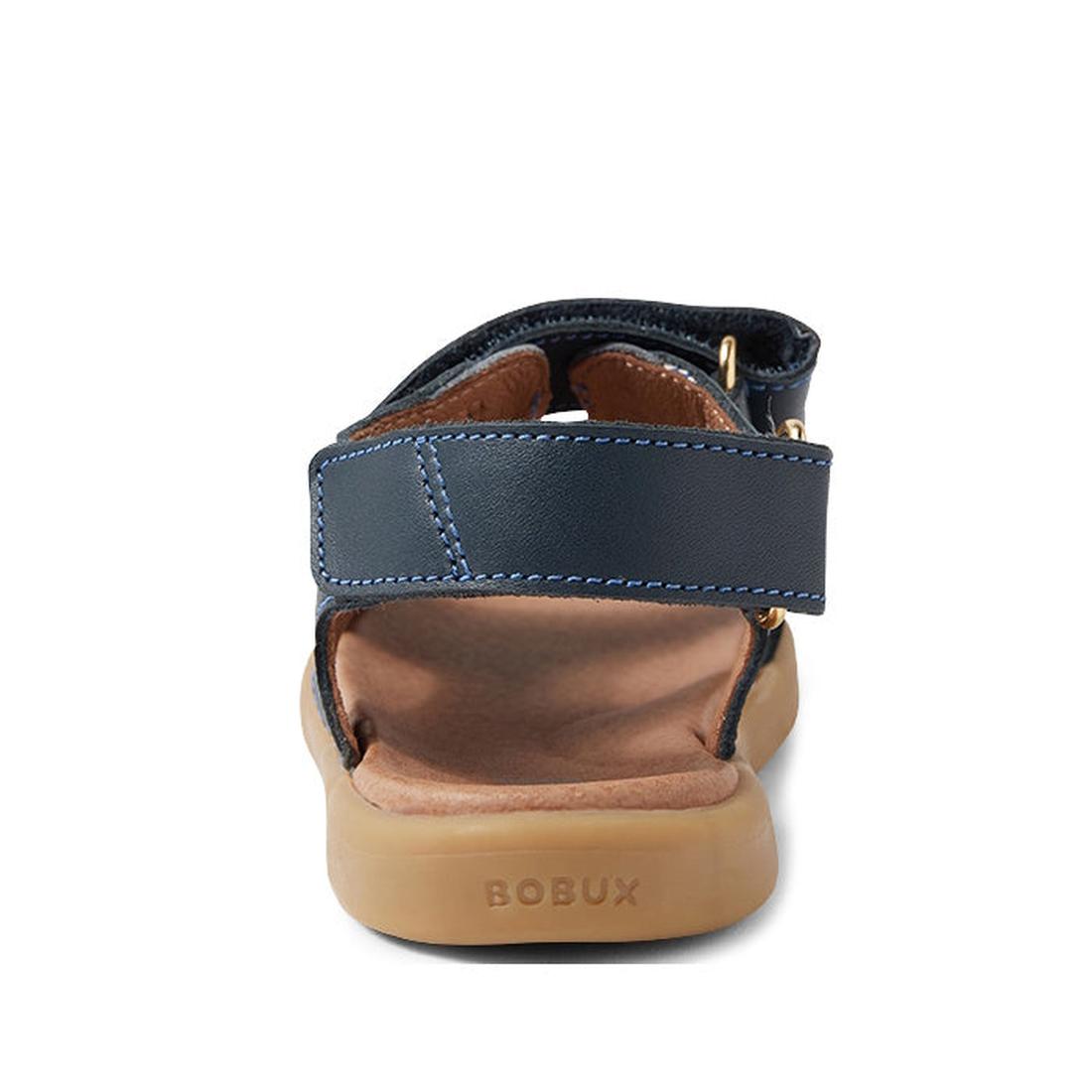 A close-up rear view of the Bobux Kid+ Plus Strand Navy Open Toe Sandal, featuring a navy blue design, brown cushioned sole with BOBUX embossed on the heel, and adjustable Velcro straps for a secure, customizable fit., 9420068557048, 9420068557055, 942006