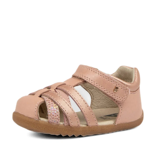 The Bobux Step Ups Cross Jump Dusk Pearl Pink Closed Toe Sandal for first walkers features a closed toe, Velcro strap, cut-out sides, and a shimmery strap detail, shown on a white background.