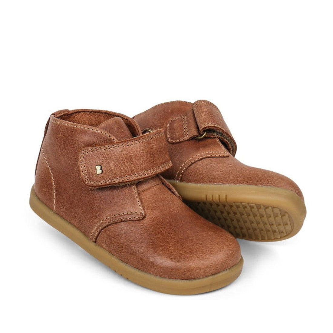 The Bobux Iwalk Desert Boot Caramel 625207A is a brown leather toddler shoe with a leather upper and velcro straps for easy wear. One upright boot and one on its side display the patterned rubber sole.