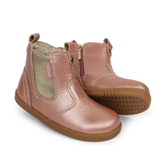 The Bobux Iwalk Jodhpur Boot Rose Gold 620839 features metallic pink leather, side zippers, beige elastic panels, pull tabs, and brown rubber soles. One boot stands upright while the other is slightly angled forward.