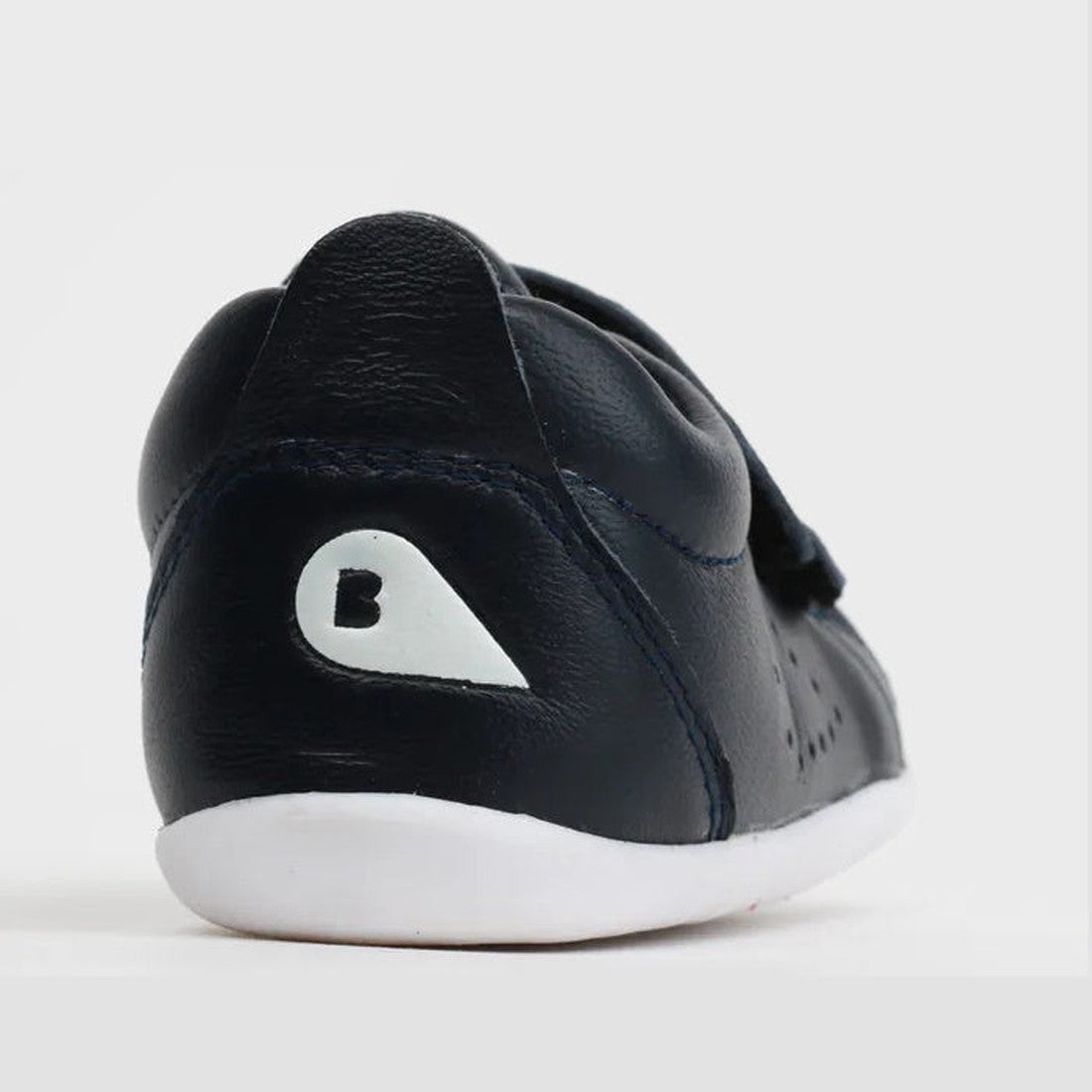 A close-up of the back of the Bobux Step Up Grass Court Trainer 728915J in navy leather, a small toddler shoe with a white sole and a white teardrop-shaped design featuring a black B on the heel., 9420062001714, 9420075792135, 9420075789753, 9420075791428