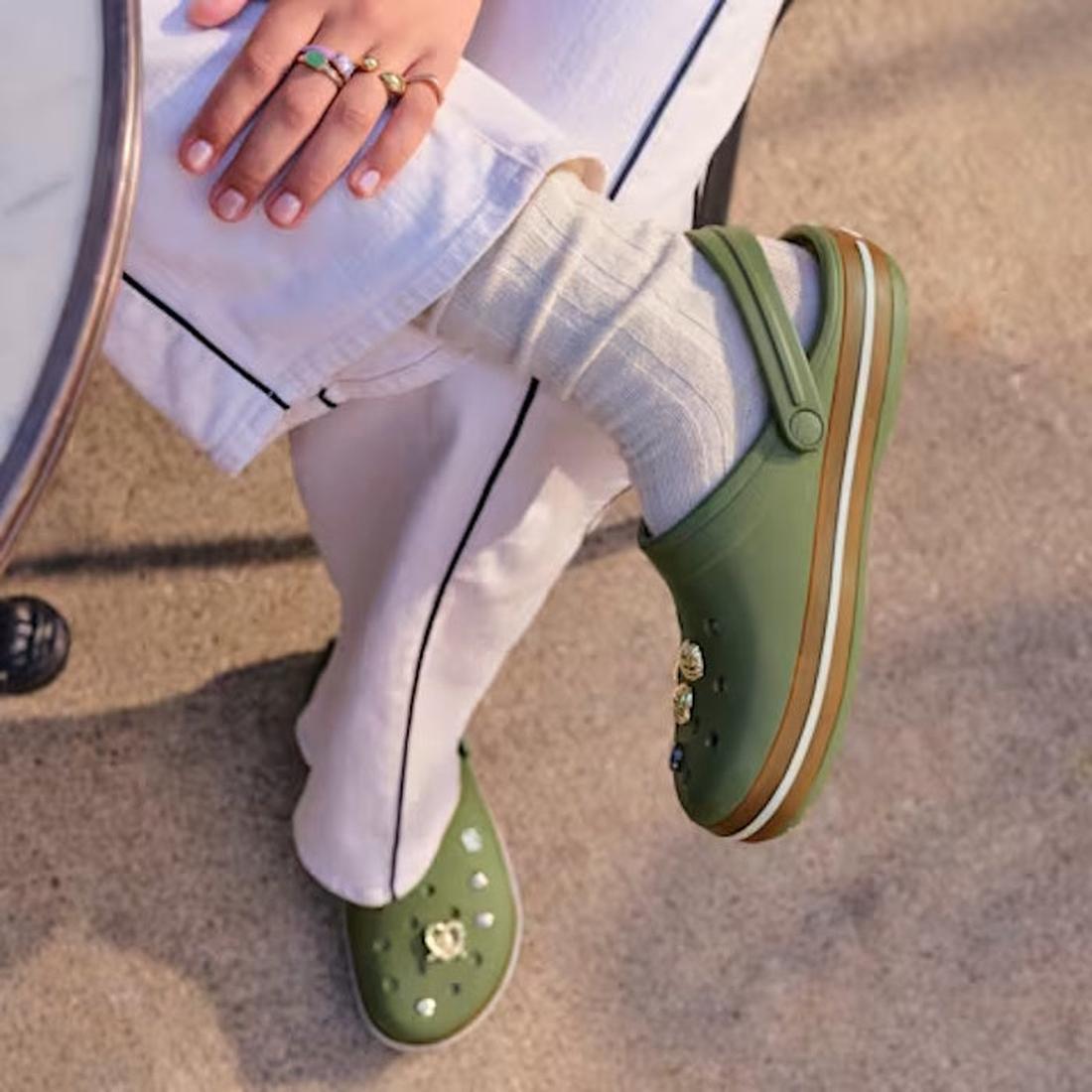 A person in cream pants with a black stripe, cream socks, and Crocs Adults Crocband Gum Clog Cargo Green with Croslite foam charms sits by a round table on sunlit pavement, their ring-adorned hand resting on their knee., 51556, 198445373183, 198445373190,