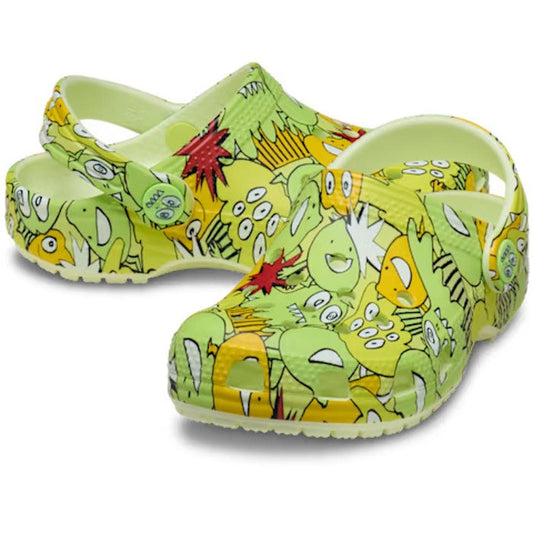 A pair of Crocs Toddlers Classic Clog Glow Monster Camo Clog Glow In the Dark, featuring a lively monster pattern with cartoon faces, teeth, and eyes in yellow, green, and red. These vibrant green clogs include a back strap and textured sole for comfort—a