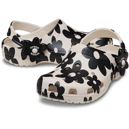 A pair of Crocs Kids Classic Clog Pop Petals Summit White Multi featuring a stylish black floral print and flower-shaped decorations around the heels. These clogs offer ventilation holes, adjustable heel straps, and provide all-day Crocs Comfort.