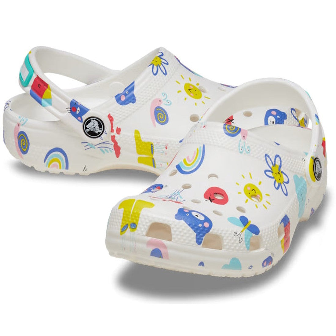 The Crocs Kids - Youths Classic Clog K Doodle Print White Multi Sandal features colorful doodles like rainbows and cartoon faces, plus ventilation holes and a heel strap—fun, customizable clogs perfect for back-to-school kids.