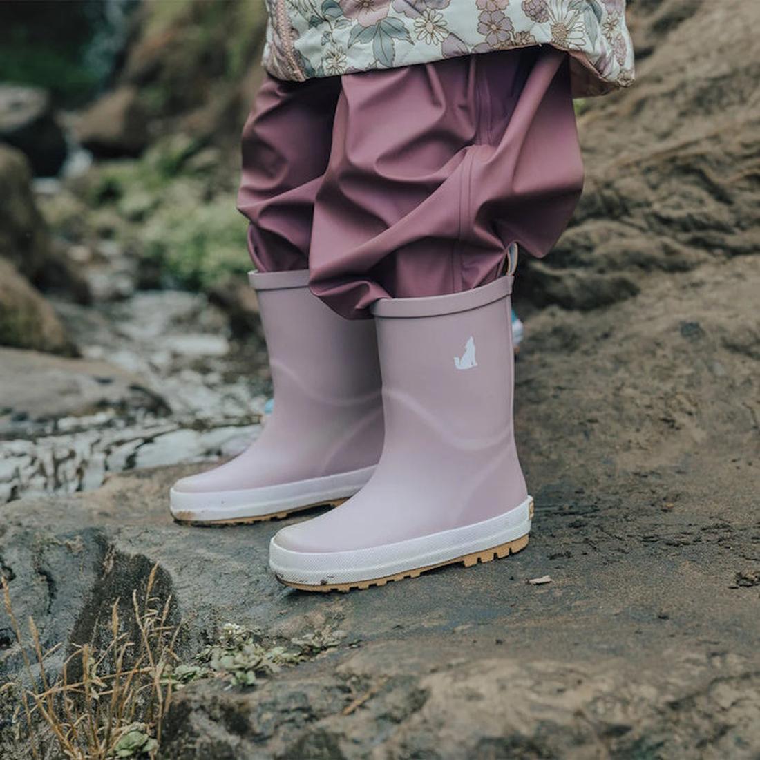 A child wearing CryWolf Rain Boots Mauve and matching waterproof pants stands on wet, rocky ground outdoors. The natural rubber boots feature a small white animal logo on the side.