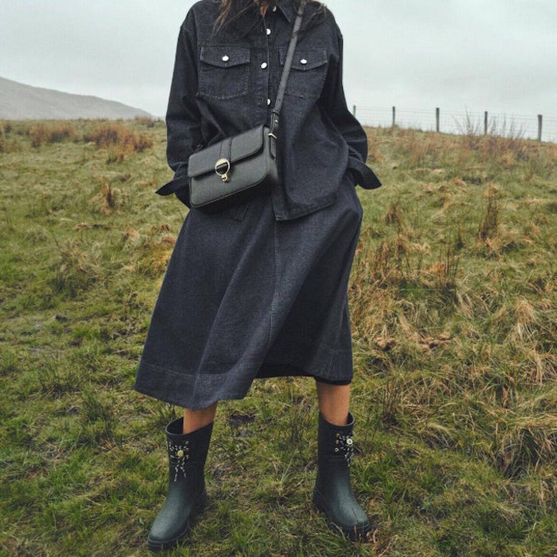 A person stands on grassy terrain under cloudy skies, wearing a dark denim shirt and midi skirt, and carrying a small crossbody bag. They complete the look with Barbour Devlin Black Mid Calf Wellys Womens Rainboot featuring floral designs., 51446, 5063669