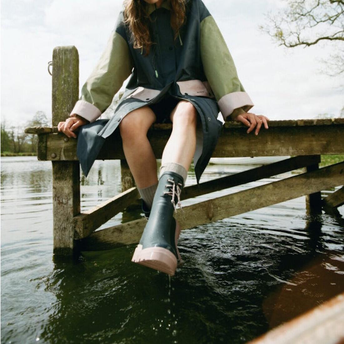 A person in a raincoat sits on a wooden dock, dipping one Barbour Devlin Dusky Grey/Gardenia Pink Mid Calf Wellys Womens Rainboot into the lake. Their face is hidden; trees line the calm, outdoorsy background., 5063669171608, 5063669171615, 5063669171622,