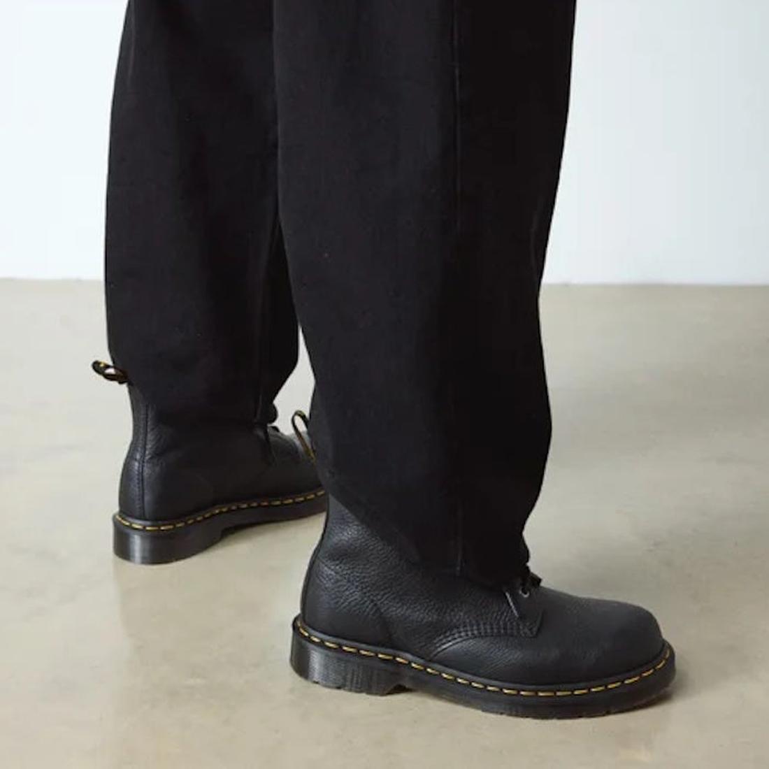 A person in black pants and Dr. Martens 101 UB Black Ambassador 6-Eye Leather Boots with yellow stitching stands on a light floor, highlighting the soft leather footwear and lower legs., 190665693683, 190665693690, 190665693706, 190665693720, 190665693737