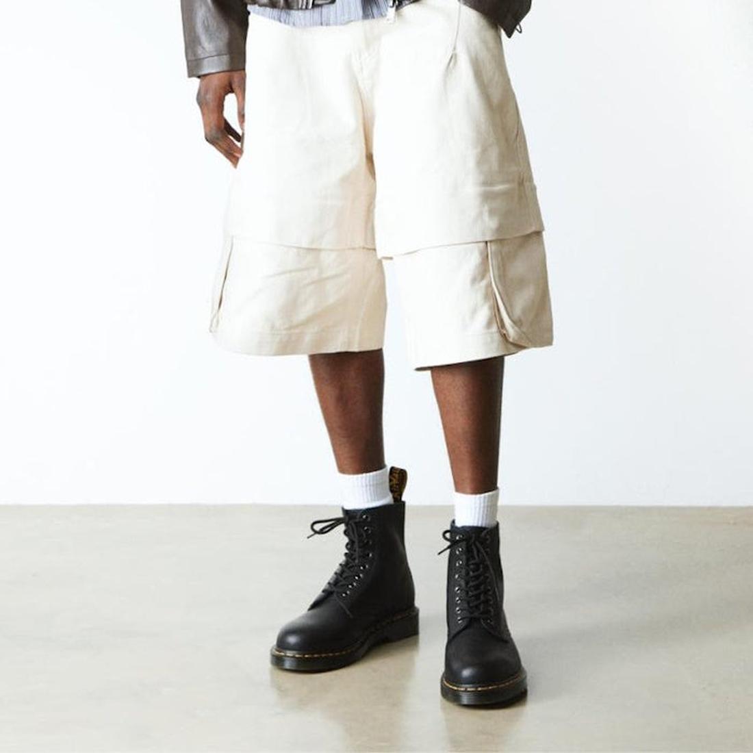 A person in cream cargo shorts, white socks, and black Dr. Martens 1460 Pascal Ambassador Leather boots stands on a light floor against a plain white background. Only the lower half of the body is visible., 46779, 190665663136, 190665663143, 190665663150,