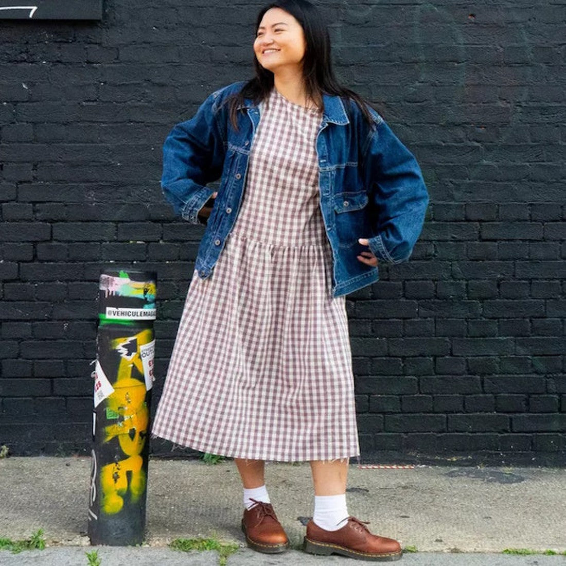 A woman smiles before a black brick wall, wearing a plaid dress, blue denim jacket, white socks, and Dr Martens 1461 Shoes in Cashew Tan Ambassador Leather. A sticker-covered pole is seen to her left.