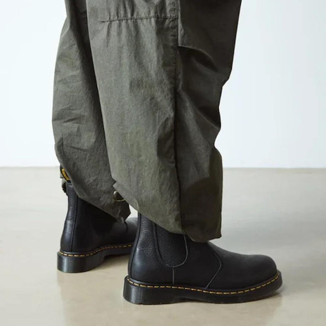 Person in loose olive green pants and Dr. Martens 2976 Ambassador Black Leather Chelsea Boots with Goodyear-welted yellow stitching, standing on a smooth, light-colored floor., 50056, 50057, 50059, 190665663303, 190665663310, 190665663327, 50067, 50068, 1