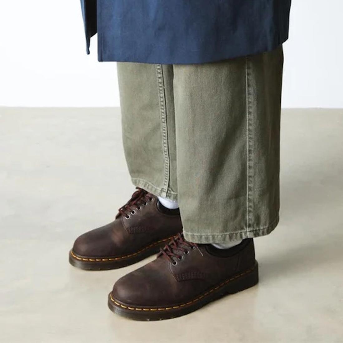 A person in wide-leg olive green pants and Dr Martens 8053 Crazy Horse Dark Brown Leather 5 Eye shoes with yellow stitching stands on a light floor. The unisex shoe has an air-cushioned sole, and the blue coat's lower part is visible., 52072, 52073, 80009