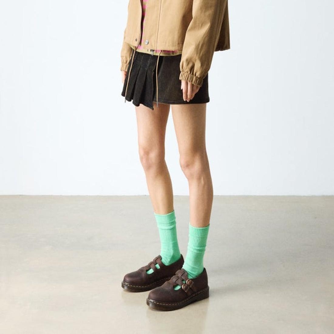 A person stands indoors on a polished concrete floor wearing a tan jacket, black pleated skirt, bright green socks, and dark brown Dr Martens 8065 2 Strap Mary Jane Shoe Crazy Horse Brown. Their face is not visible., 190665588941, 190665588958, 1906655889
