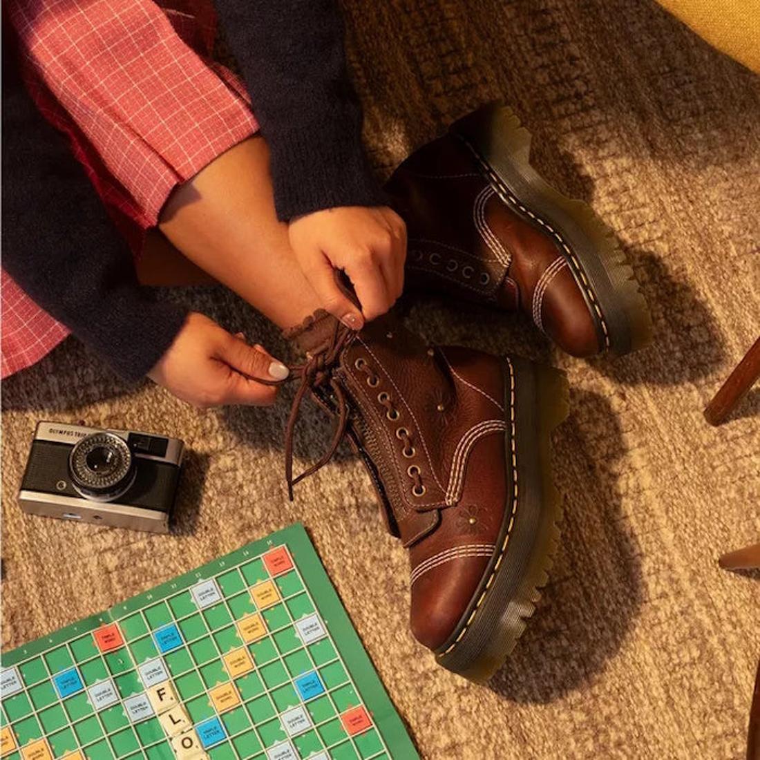 Someone wearing pink plaid pants and Dr Martens Sinclair Flower Dark Brown Grizzly Leather Boots with a platform Quad sole ties their laces beside a vintage camera and Scrabble board on a carpeted floor., 51903, 51904, 190665777116, 190665777123, 19066577