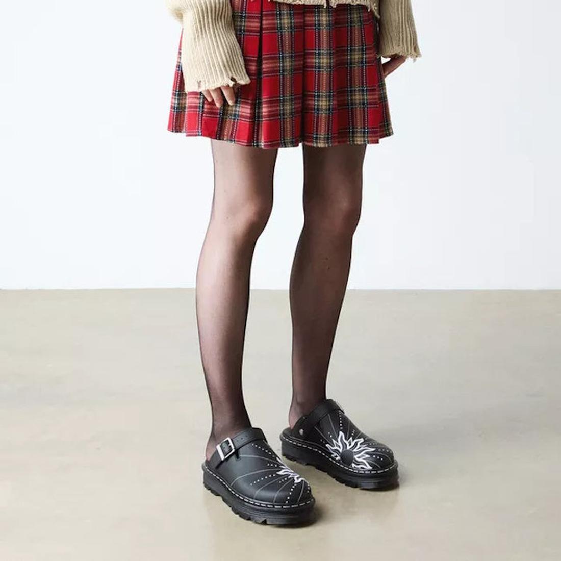A person in a red plaid skirt, sheer black tights, and Dr Martens Zebzag Mule Solar Flare Genix Nappa Black clogs stands on a smooth, light-colored floor. Only the lower body is visible., 190665758818, 190665758825, 190665758832, 190665758856, 19066575886