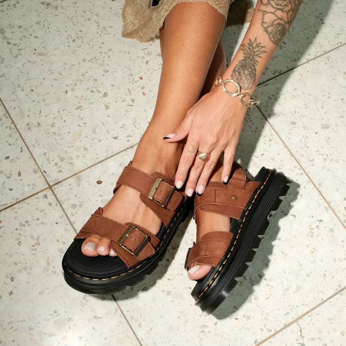 A person with a tattooed arm wears Dr Martens Zebzag Sepia Brown Nubuck 2 Strap Sandals with black soles and gold buckles, sitting on a speckled tile floor. Their ring-and-bracelet adorned hand rests on their foot., 190665790603, 190665790610, 19066579062