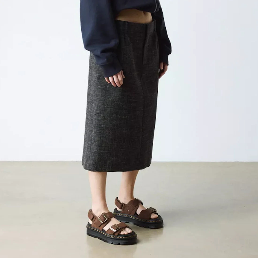 Indoors, a person wears a dark gray knee-length skirt, navy long-sleeve top with extended sleeves, and Dr Martens Zebzag Sepia Brown Nubuck 2 Strap Sandals. The minimal background features a light floor and wall., 190665790603, 190665790610, 190665790627,