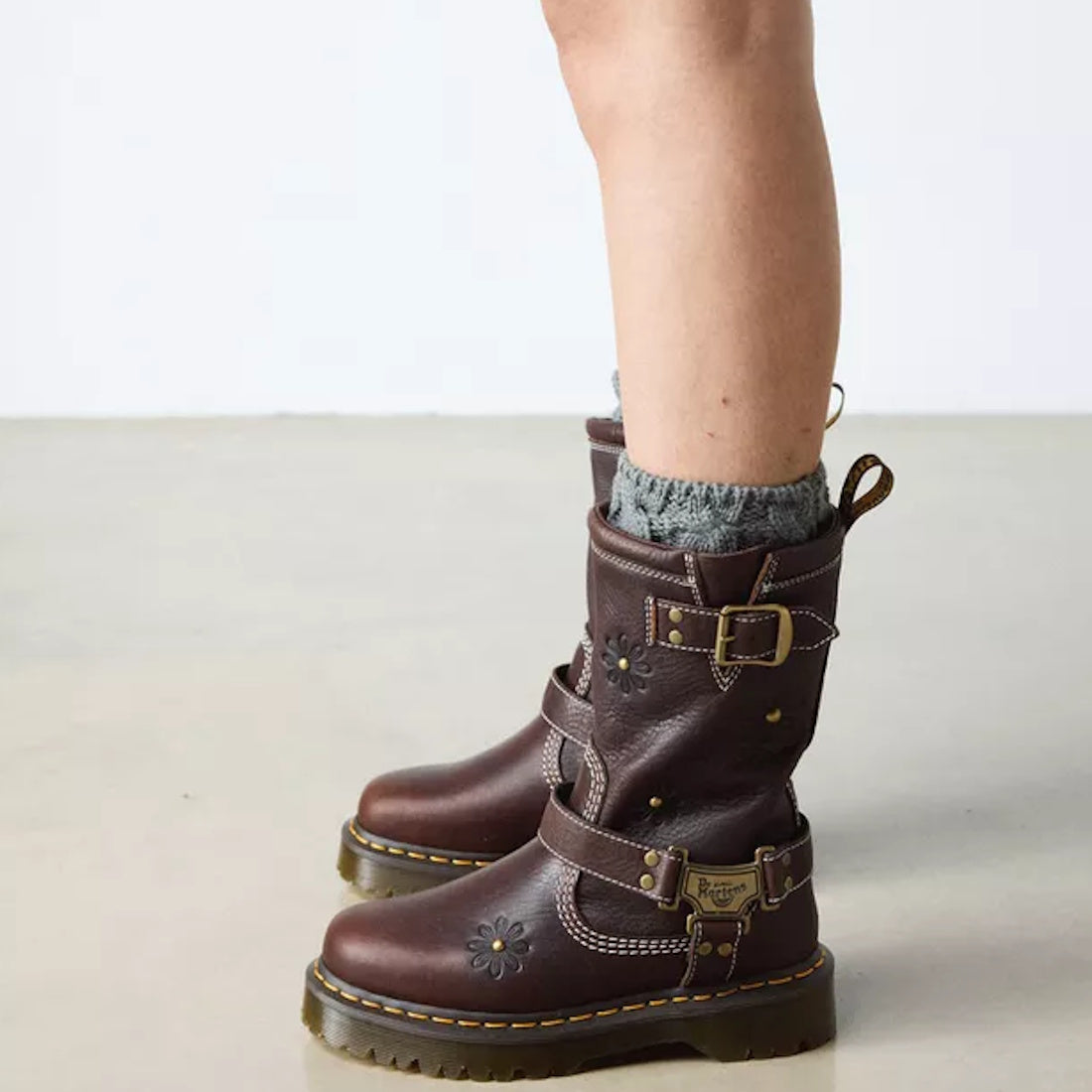 Someone wearing gray knit socks and Dr Martens Anistone Hi Flower Bex Grizzly Dark Brown boots, featuring buckles, decorative stitching, and floral details, stands on a light floor with a plain background.