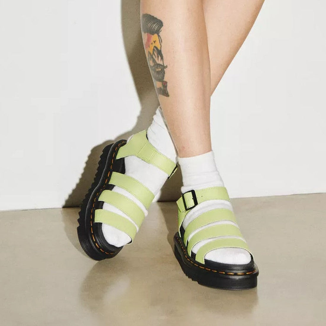 A person with a calf tattoo wears white socks and Dr Martens Blaire Lime Green Athena 3 Strap Sandals with black soles and yellow stitching, standing on a beige floor against a white wall., 190665644951, 190665644968, 190665644975, 190665644999, 41506, 41
