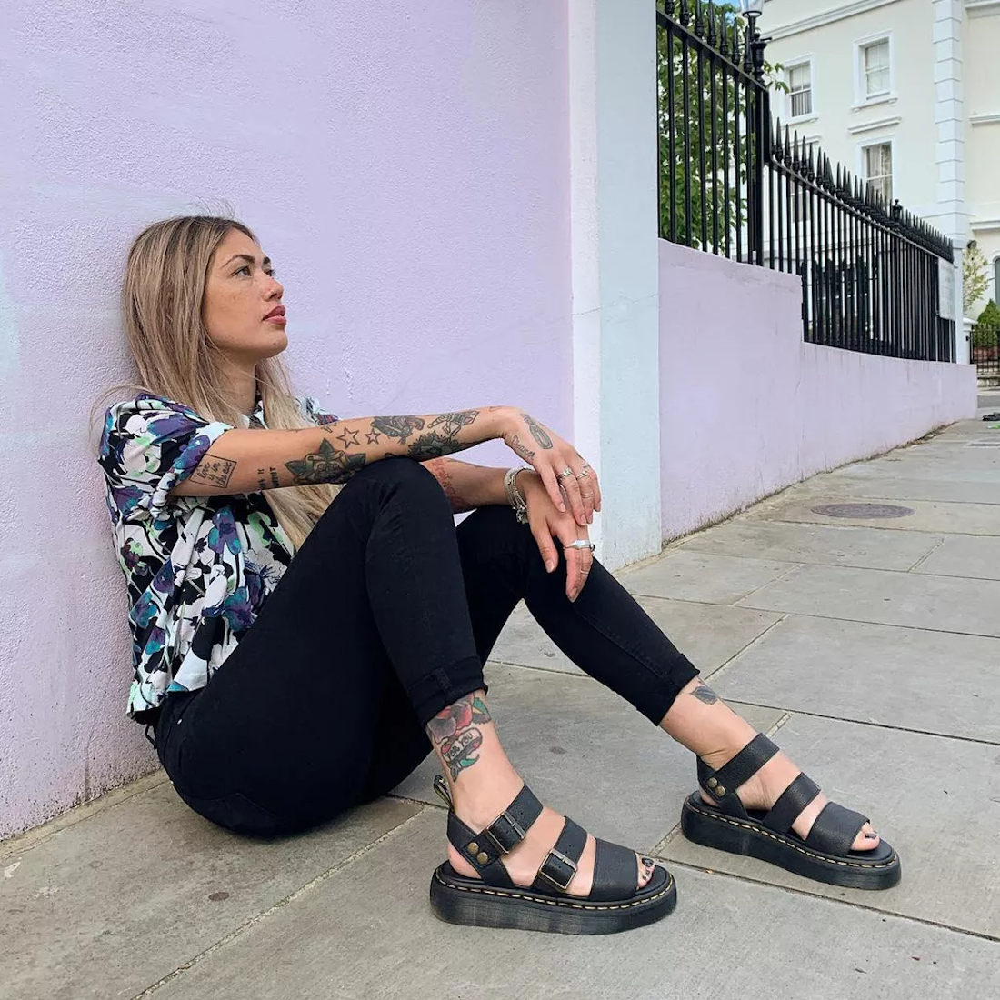 Woman sitting on a sidewalk wearing  Dr Martens Black Gyphon black sandals in pisa leather and a colorful shirt against a light purple wall.,
190665318654,
190665318661,
190665318678,
190665318685,
190665318692,
190665318708,
190665318715,, 190665318654, 