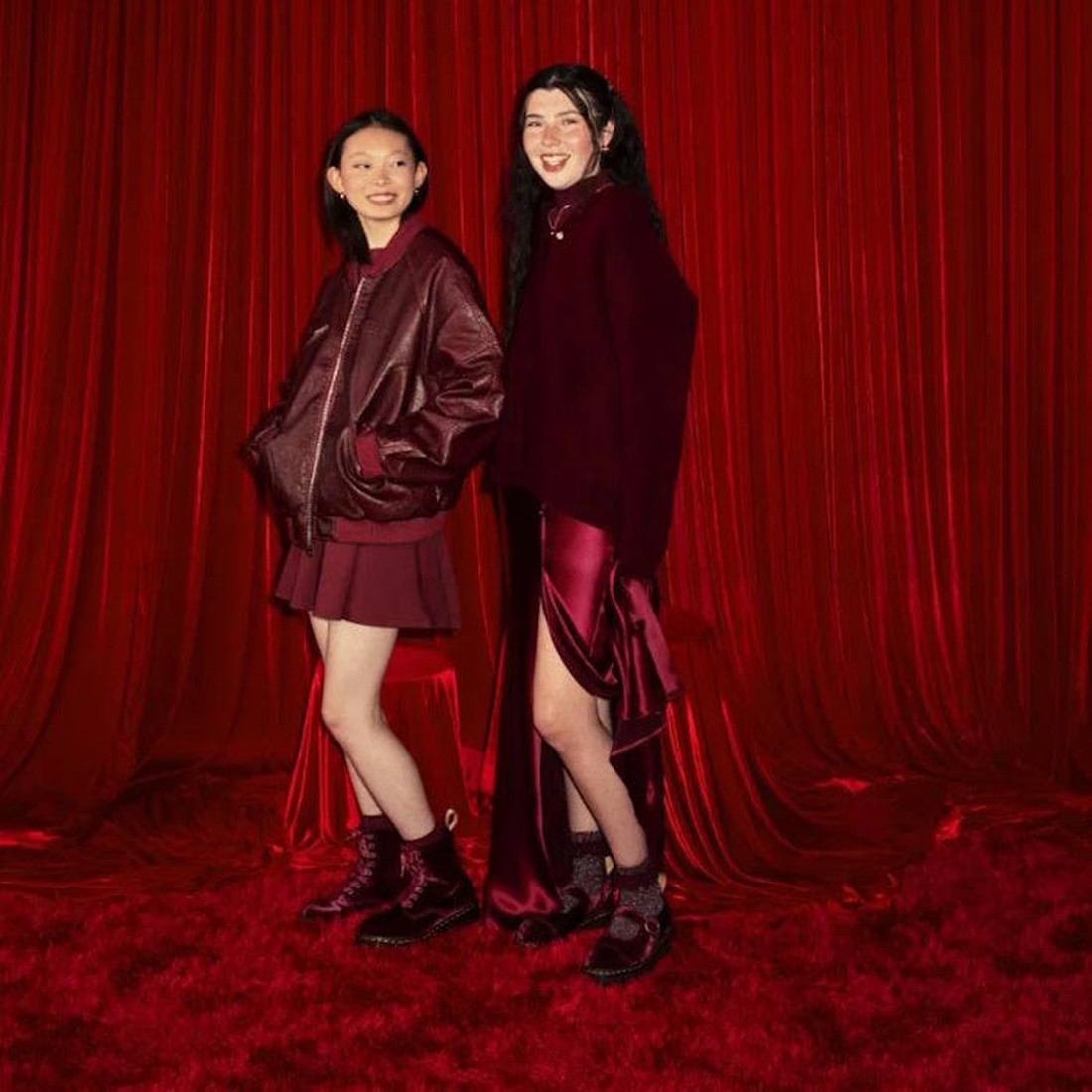 Two young women smile in front of rich red curtains and carpet, both dressed in chic burgundy layers and Dr Martens 1460 Vegan Pascal Cherry Red Velvet 8 Eye Boots, perfectly complementing the vibrant, grunge-inspired backdrop., 42020, 190665660616, 19066