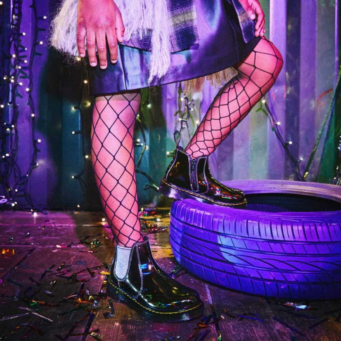 A person stands with one foot on a purple tire, wearing a shiny dark skirt, fishnet tights, and Dr Martens Kids Juniors 2976 J Chelsea Boot Black Lightshow Sparkle Rays. Colorful lights and confetti create a festive scene., 190665495171, 190665495195, 190