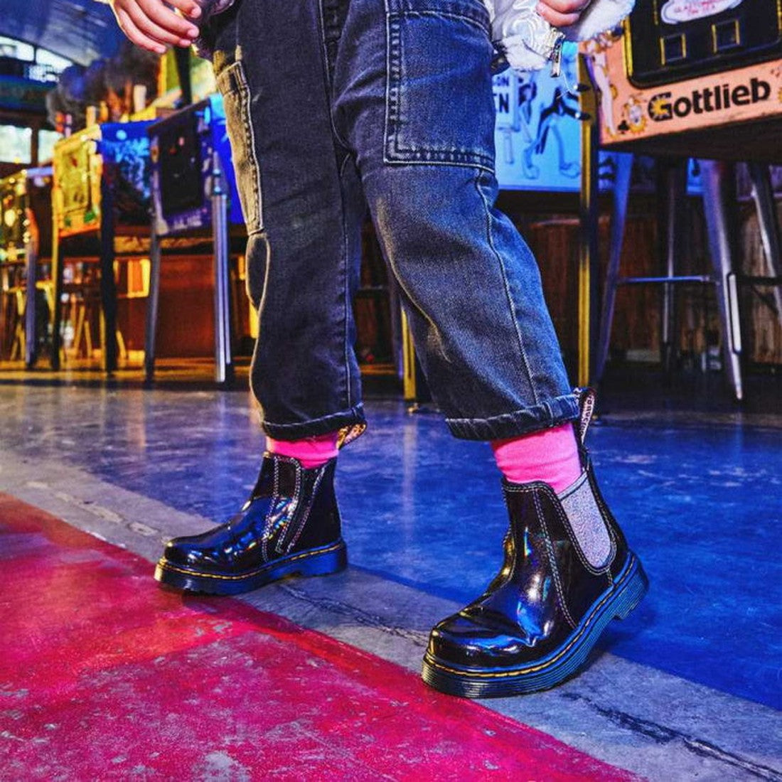 A person in cuffed jeans and bright pink socks stands on a shiny blue and red floor, wearing Dr Martens Kids Toddlers 2976 T Chelsea Boot Black Lightshow Sparkle Rays (toddlers UK8), with arcade games and colorful lights in the background., 190665494914, 