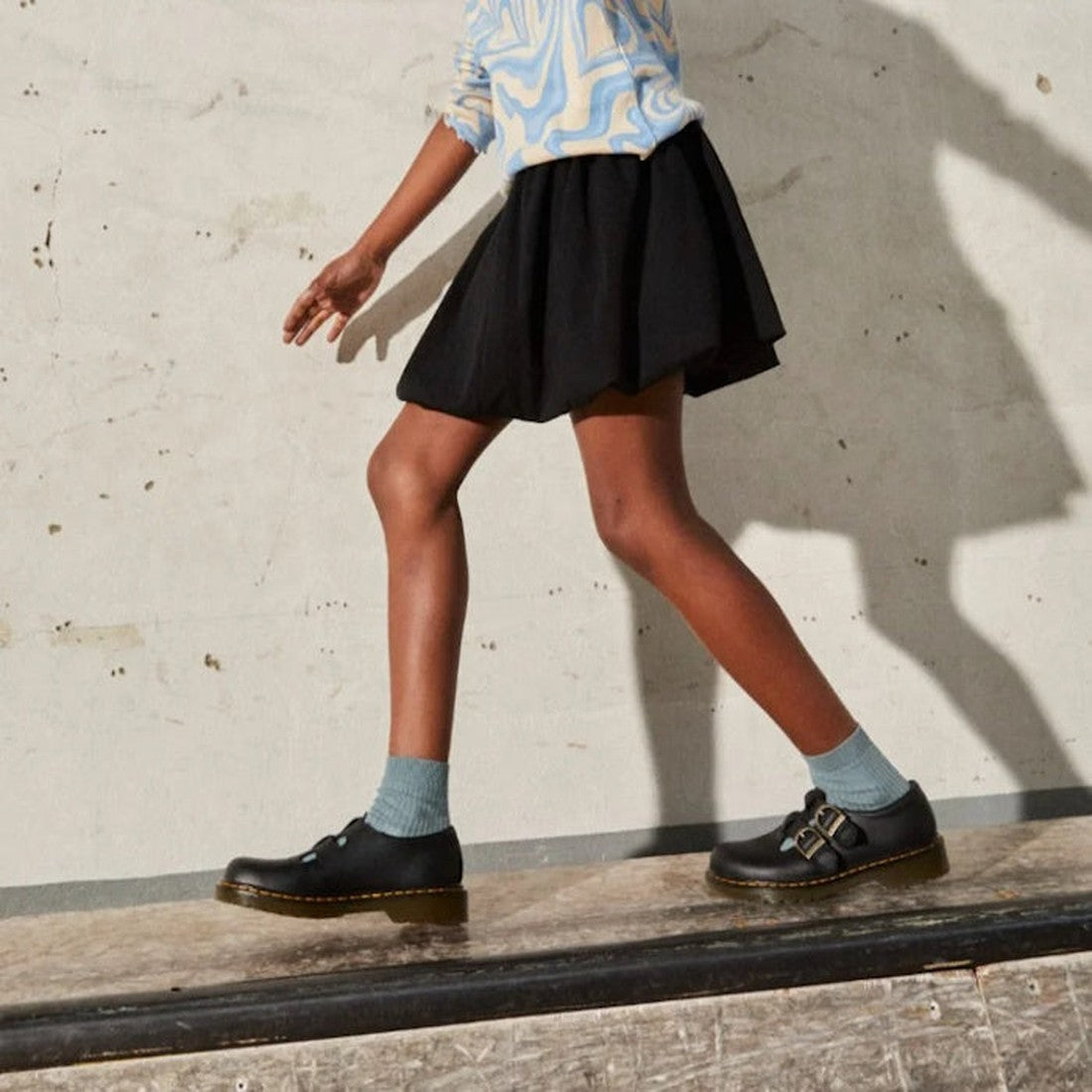 A person’s lower half is visible, showing them walking on concrete in a patterned shirt, black skirt, blue socks, and Dr Martens Kids 8065 J Black Softy T Mary Jane Leather Shoes with an air-cushioned sole., 41982, 41983, 190665537093, 190665537109, 19066