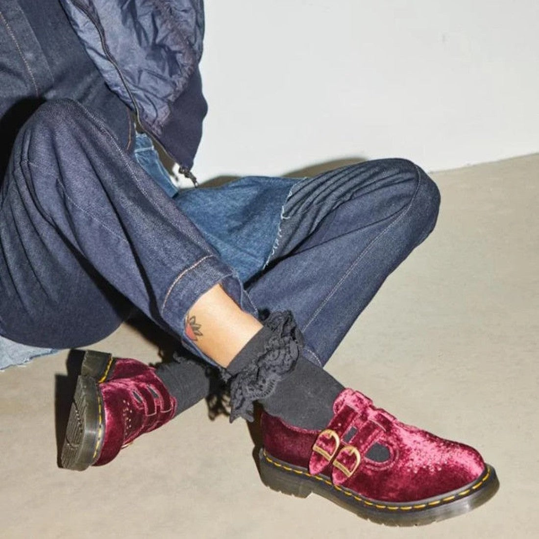 A person sits on the floor, lower half visible, wearing dark denim jeans, a jacket, black ruffled socks, and Dr Martens 8065 2 Strap Mary Jane Vegan Cherry Red Velvet shoes with gold buckles., 42014, 190665660548, 190665660555, 190665660579, 42018, 42019
