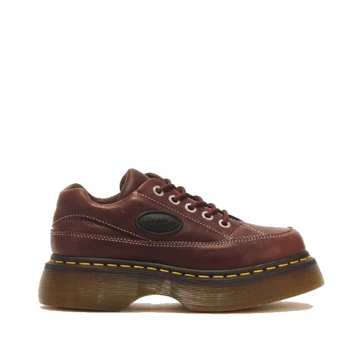 The Dr Martens Buzz 5i Dark Brown Grizzly Leather Shoe features brown leather, white stitching, metal eyelets, a thick translucent chunky barrel-edge Buzz sole with yellow stitching, and a black oval logo patch on the side, bringing nostalgic 90s silhouet