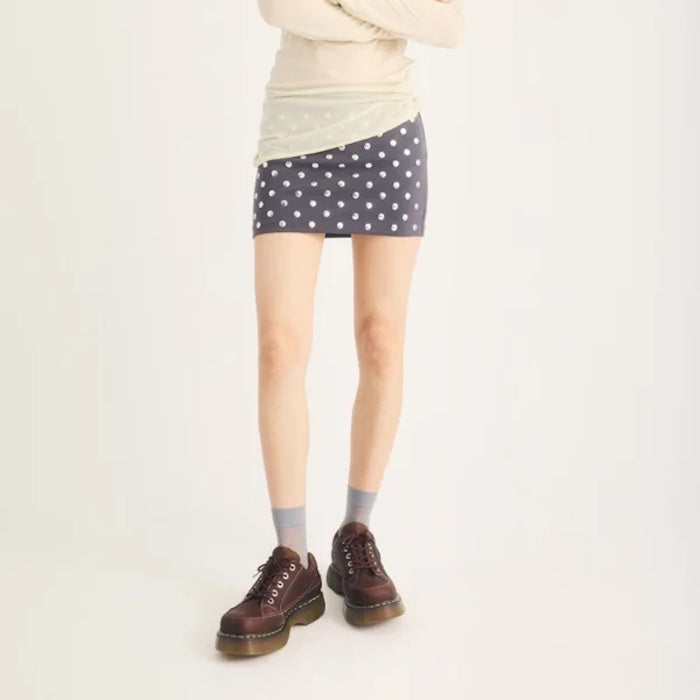 A person stands against a plain background with their arms crossed and face not visible, wearing a beige long-sleeve top, a blue polka dot mini skirt, gray socks, and Dr Martens Buzz 5i Dark Brown Grizzly Leather Shoes featuring brown platform oxford styl