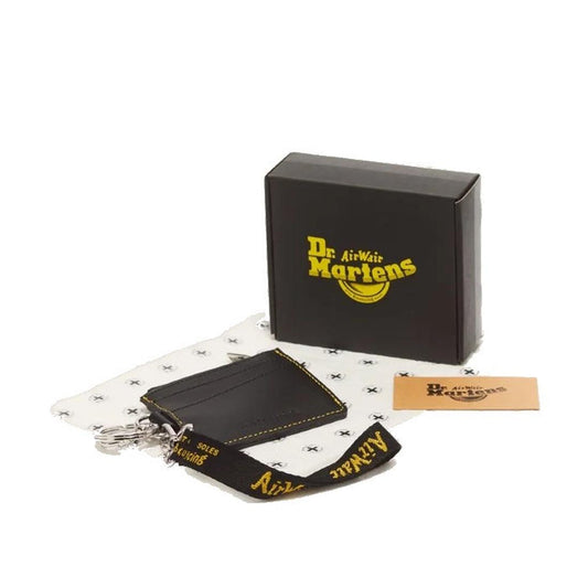 Displayed on a white background are a black Dr. Martens box, a brown card, and the Dr. Martens Cardholder Keyring Black Kiev Leather Bag Charm with a white cloth. The black leather cardholder features yellow Dr. Martens branding on its detachable lanyard,