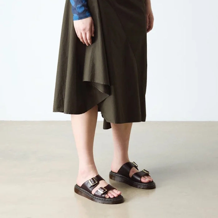 A person wears a dark olive asymmetrical midi skirt and black Dr Martens Josef Charro Atlas Slide Sandal Leather on a light floor, with only the lower half of their body visible.