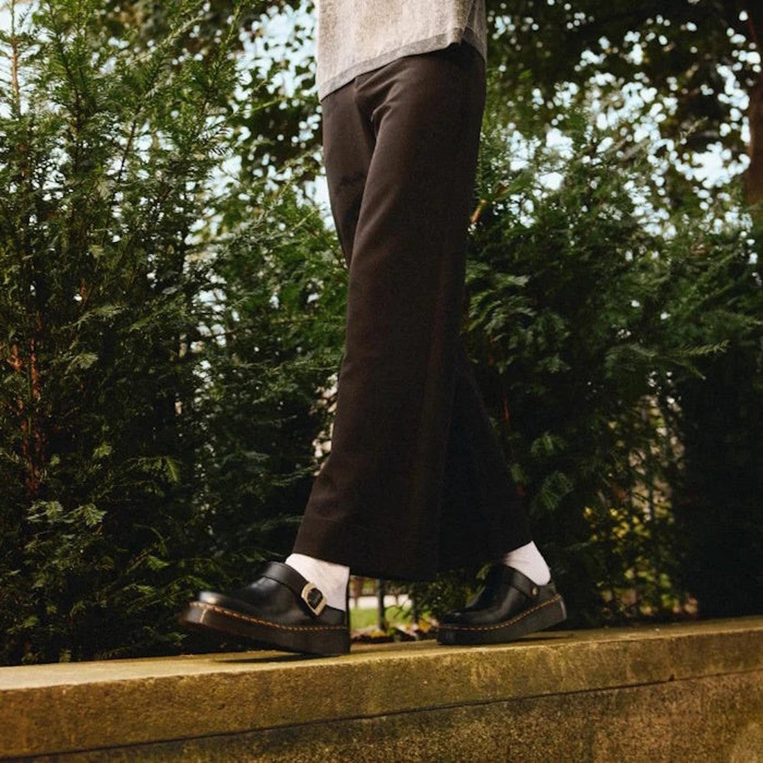 A person in black pants, white socks, and Dr Martens Laketen Black Atlas Archive Mule Slide Sandals stands on a low ledge outside, with green bushes and trees in the background., 190665644302, 190665644319, 190665644333, 190665644340, 190665644357