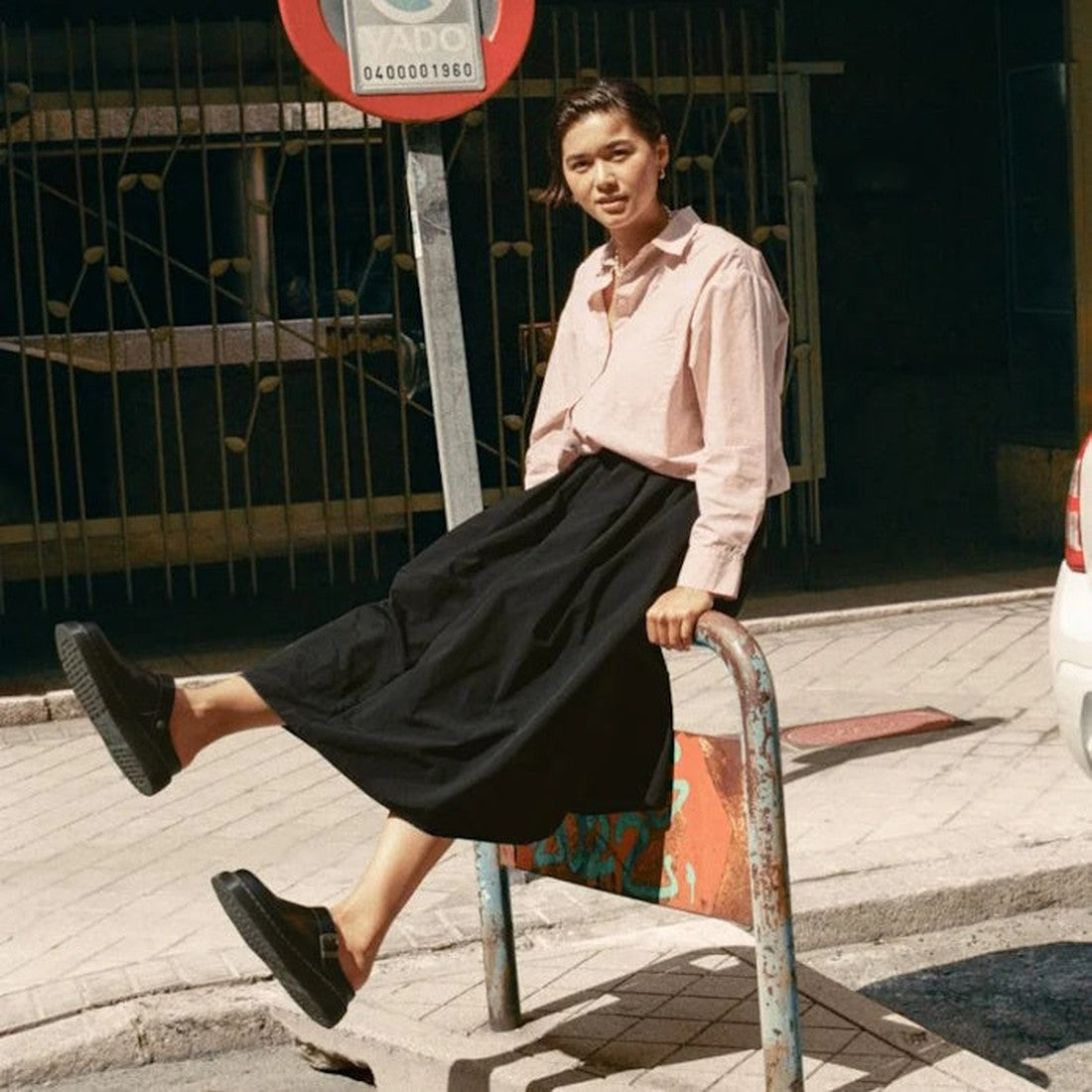 A woman in a pink shirt, black skirt, and Dr Martens Laketen Black Atlas Archive Mule Slide Sandal sits casually on a metal rail along a sunny sidewalk, with buildings and parked cars in the background., 190665644302, 190665644319, 190665644333, 190665644340, 190665644357