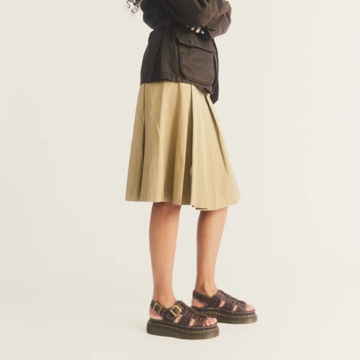 A person wearing a tan pleated skirt and Dr Martens Wrenlie Flower Open Toe Dark Brown Grizzly Quad Sandal, featuring floral cutouts and a Quad Shore sole.