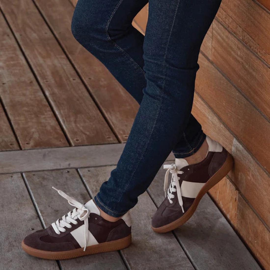 Wearing dark jeans and EOS Antigo Cacao Brown Leather Lace Up Trainers, featuring white stripes and gum soles, standing on a wooden floor and leaning against a wooden wall. Only legs and shoes are visible., 51643, 9321191255156, 9321191255163, 93211912551