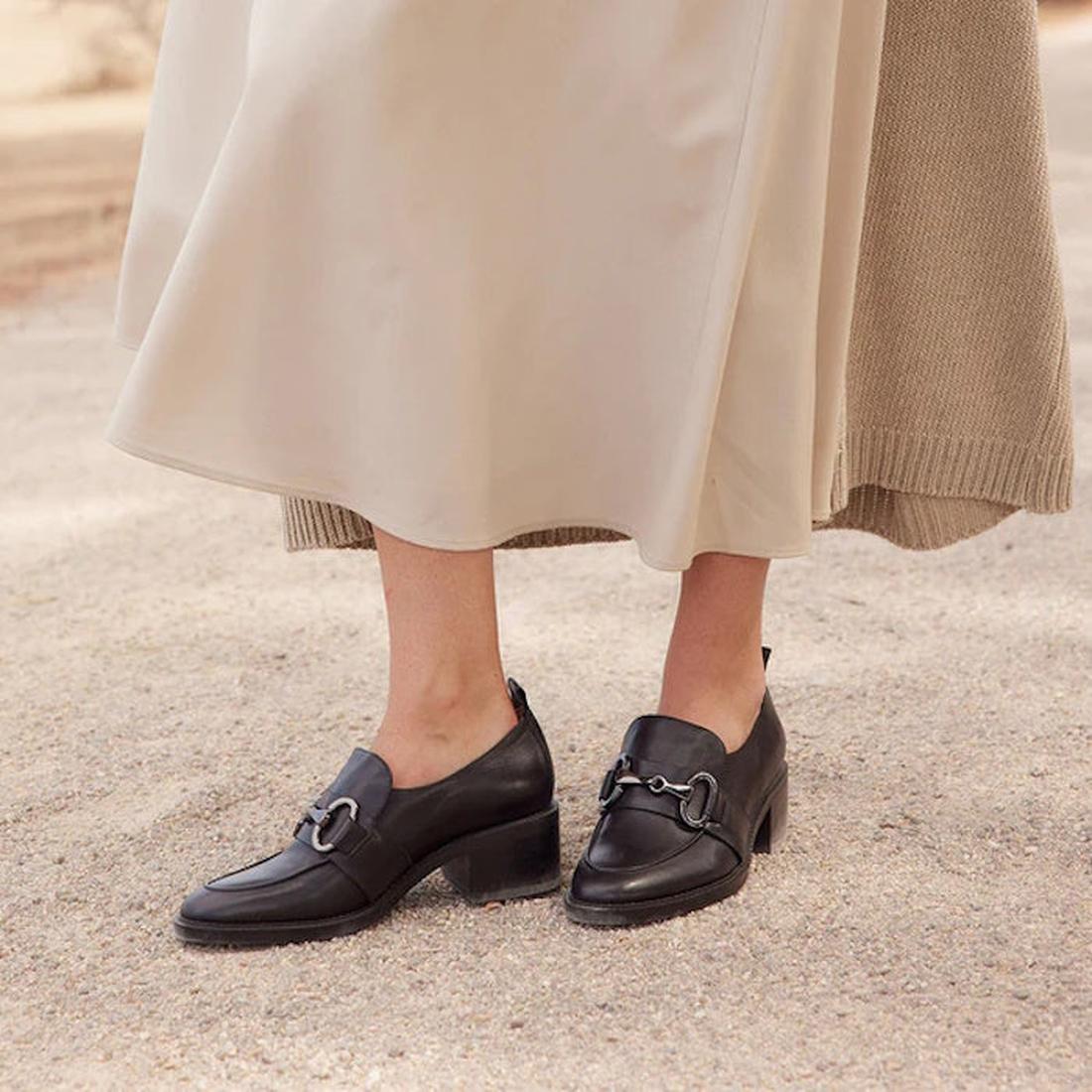 Black loafers with a slight heel on a womens feet worn with a beige skirt on a neutral background,
9321191193403,
9321191193410,
9321191193427,
9321191193434,, 45648, 9321191193403, 9321191193410, 9321191193427, 9321191193434, 45653, 45654