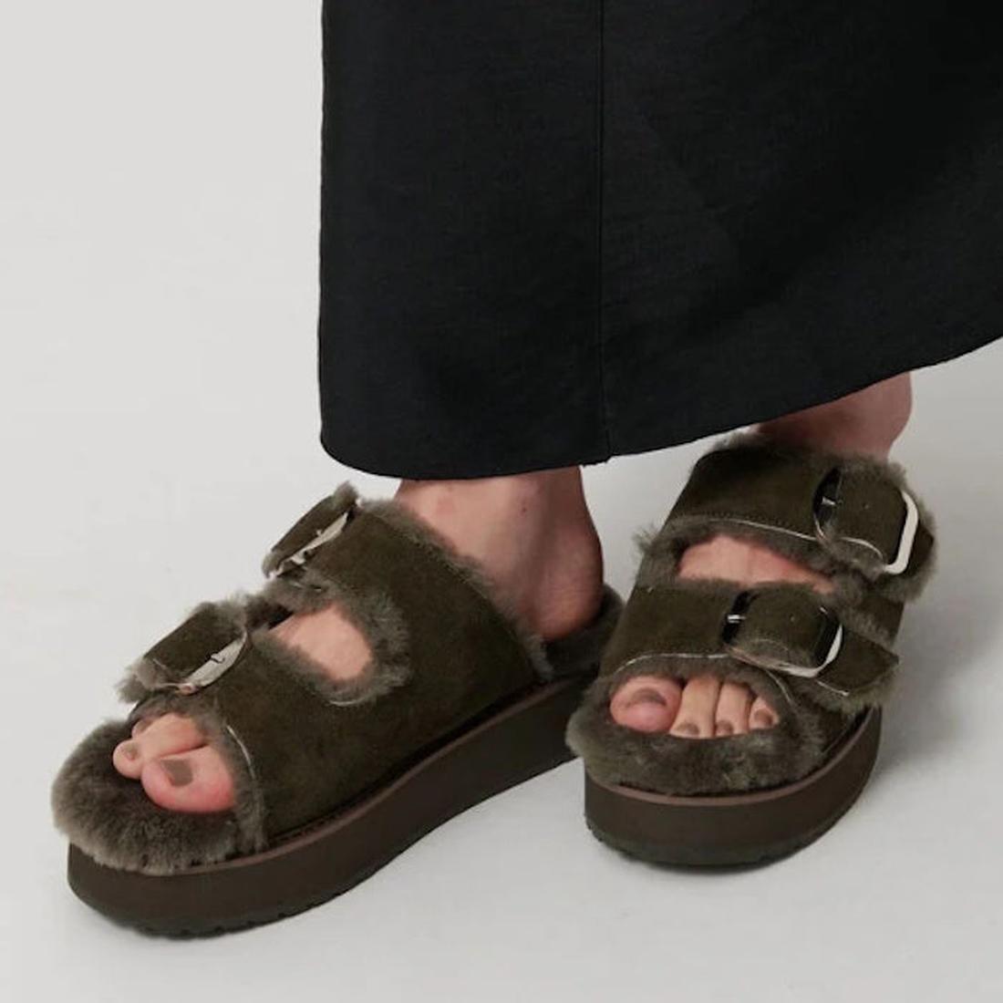 A person wears Emu Womens Cabello Dark Olive Sheepskin Wool Slipper Slides—fuzzy, two-strap slip-ons with thick soles—paired with a long black skirt., 50698, 9330071990681, 9330071990698, 9330071990704, 50702, 50703