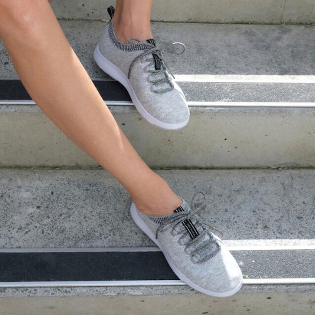 A person sits on concrete steps wearing Emu Heidelberg Wool Sneaker Grey, featuring light grey knit uppers, white soles, black and grey laces, and a black pull tab. Only their lower legs are visible., 42766, 42767, 9330071956397, 9330071956403, 9330071956