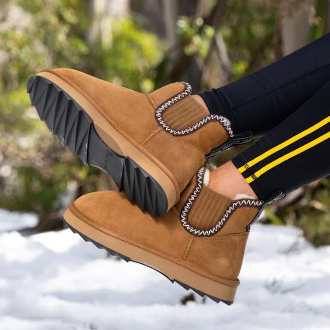 A person wearing Emu Womens Higgie Chestnut Platform Wool Boots with a chunky sole and zigzag stitching sits outdoors, surrounded by snow and greenery., 9330071978580, 9330071978597, 50686