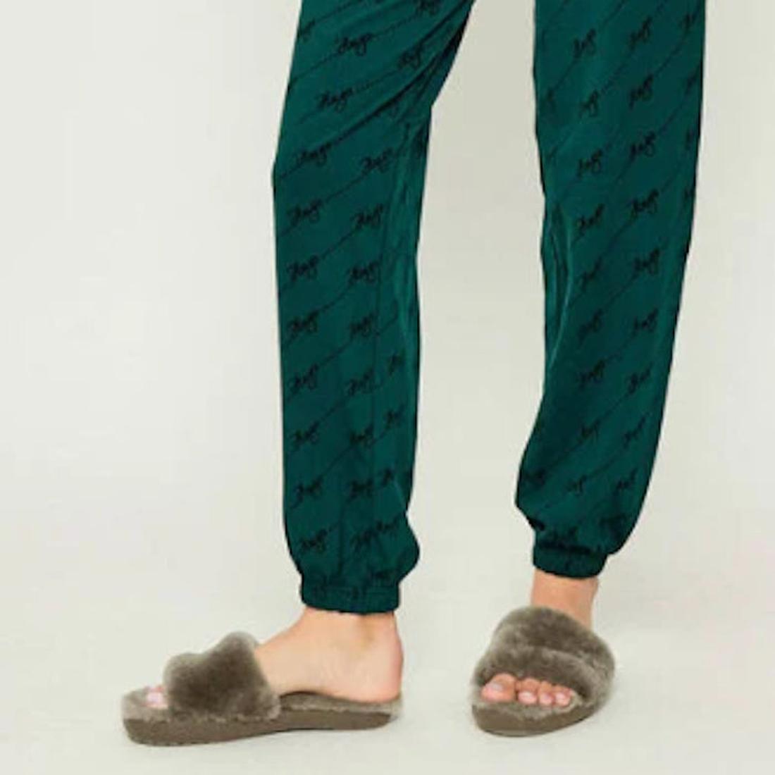 A person wearing green pajama pants stands on a light-colored floor, with only their lower legs and feet visible. They are wearing EMU Women's Mckay Dark Olive Sheepskin Scuff Slipper Slides., 50733, 9330071976265, 9330071976272, 9330071976289, 50737, 507