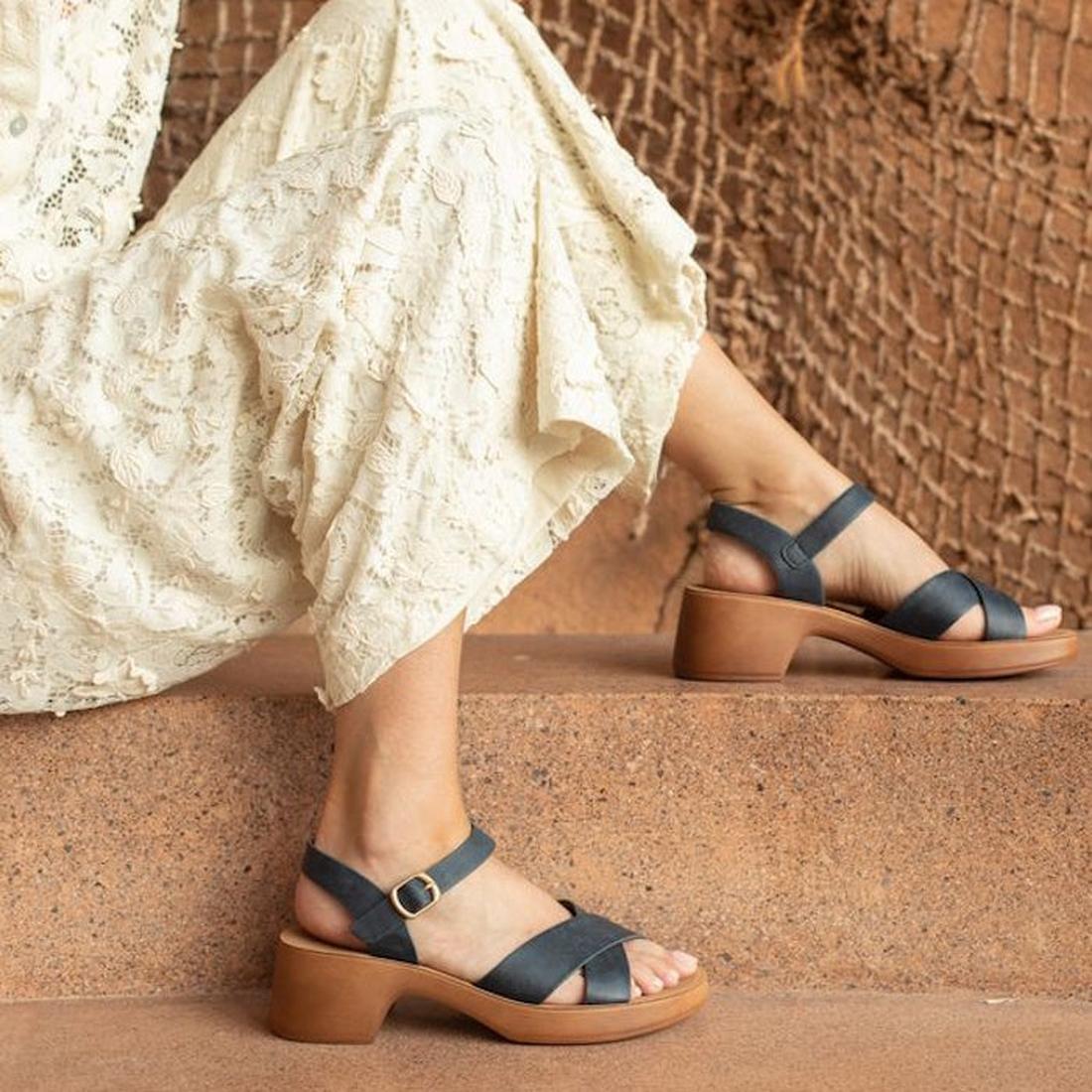 A person in a cream lace dress sits on a stone ledge, showcasing the Los Cabos Alegra Navy Wedge Platform Backstrap Sandal. A rustic net and textured brown wall provide a serene backdrop for the navy sandals with tan soles., 9321191292311, 9321191292328, 