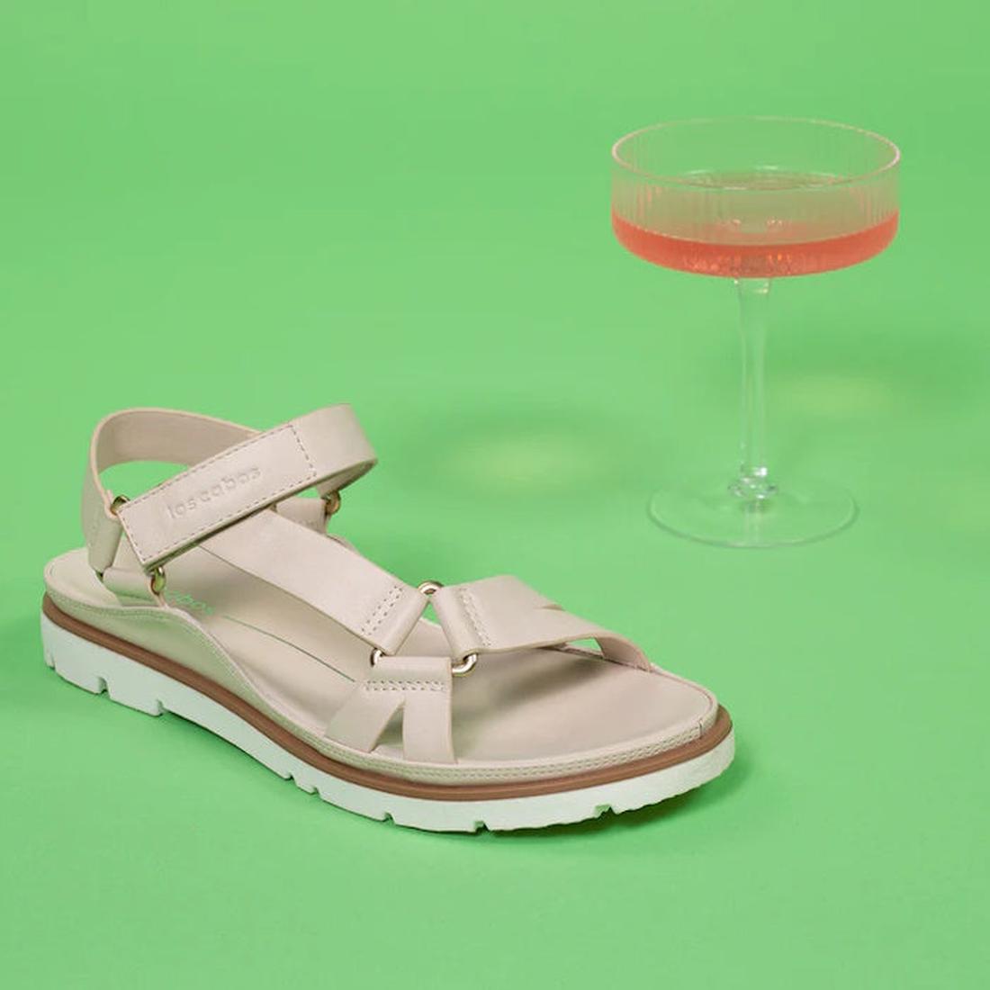 A Los Cabos Ashanti Bone Women's Sandal with back strap, cushioned footbed, and white sole is shown on a green surface beside a coupe glass filled with pink liquid., 9321191293981, 9321191293998, 9321191294001, 9321191294018, 9321191294025, 9321191294032,