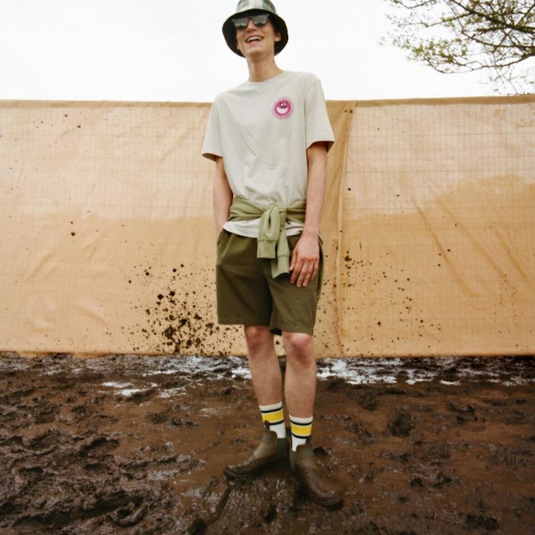 A person stands smiling on muddy ground, wearing a bucket hat, sunglasses, a beige t-shirt with a pink smiley logo, green shorts, patterned socks, Barbour Nimbus Olive/Ochre Chelsea Wellingtons Men's Rainboots, and a sweatshirt tied around their waist., 5