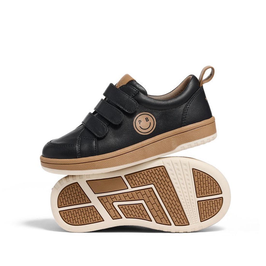 The Pretty Brave Happy Smiley Trainer Black Leather features black kids’ sneakers with tan soles, triple Velcro straps, and a smiley face PB logo on the side; one shoe stands upright while the other balances sideways underneath.
