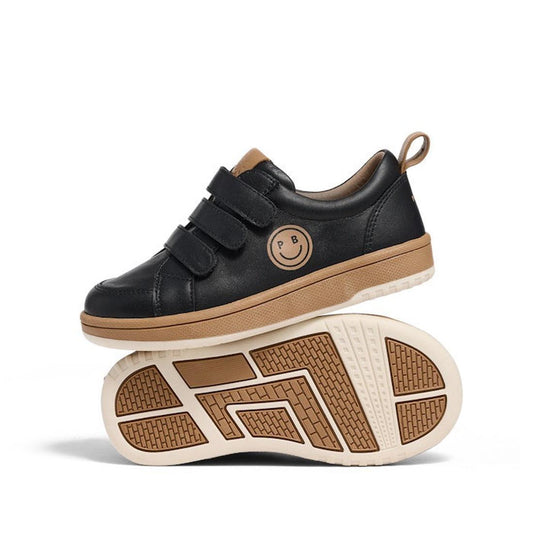 The Pretty Brave Happy Smiley Trainer Black Leather features black kids’ sneakers with tan soles, triple Velcro straps, and a smiley face PB logo on the side; one shoe stands upright while the other balances sideways underneath.