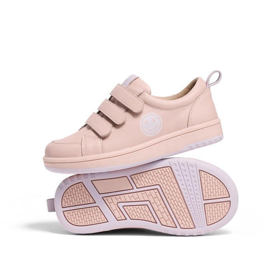 The Pretty Brave Happy Trainer Blush Pink Leather features triple velcro straps and a smiley face logo; one kids' sneaker stands upright while the other is sole-up, showing its textured tread.